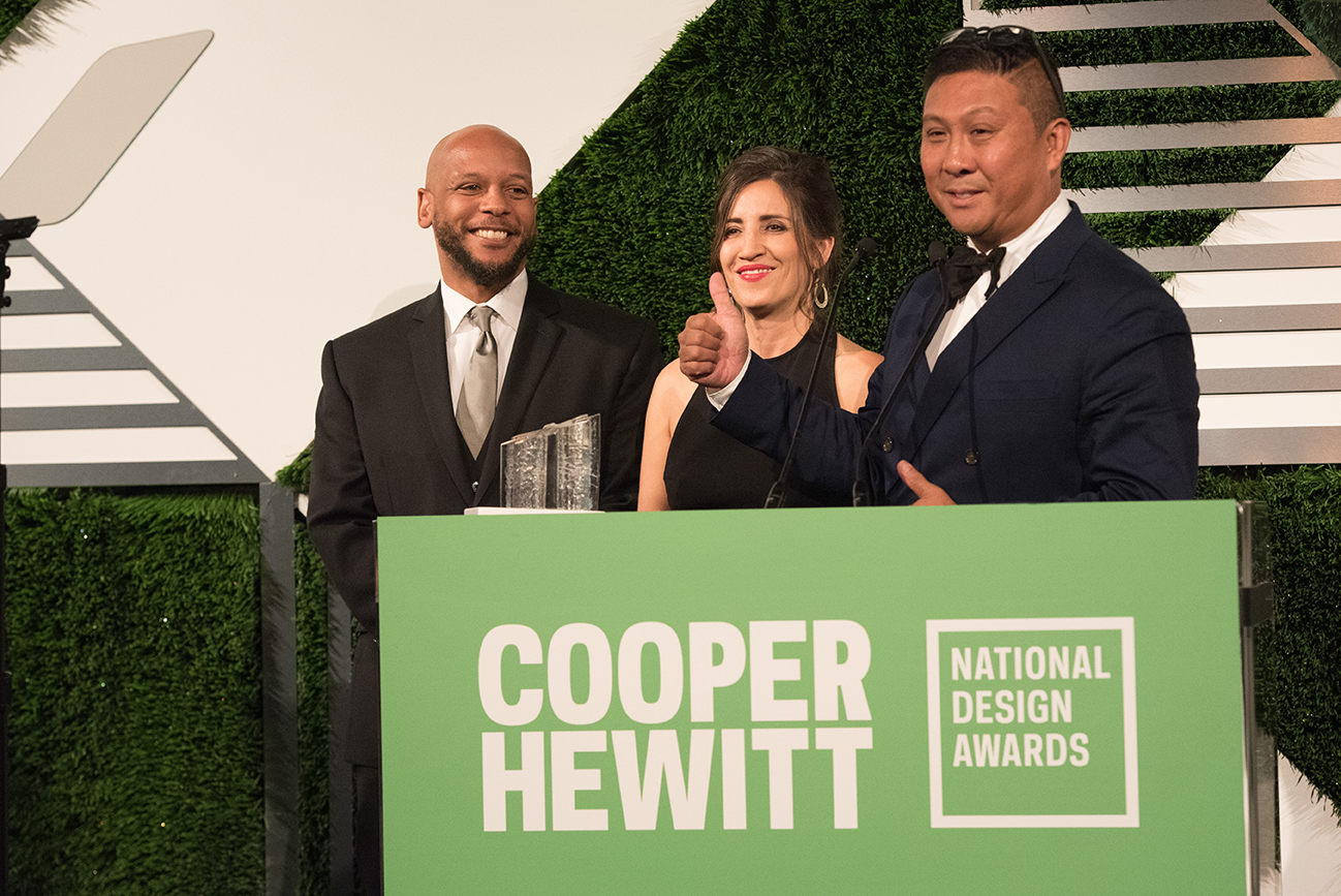 Three people stand triumphantly behind a podium at the National Design Awards. One has a hand in the