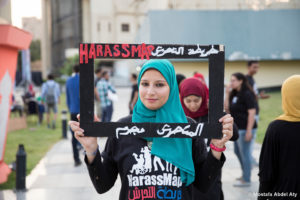 Medium-skinned woman wearing teal hijab standing outside busy pedestrian area, holding rectangular frame around her head with written red, bold text [Harassmap] and white Arabic text.