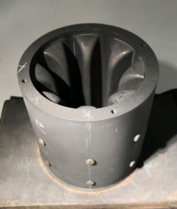 A gray cylindrical vessel with screws and white chalk marks on the outside and eight deep grooves on the inside.