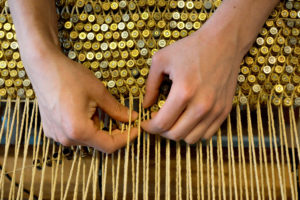 Close-up photograph of a pair of hands weaving golden bullets into a loom stretched with twine.