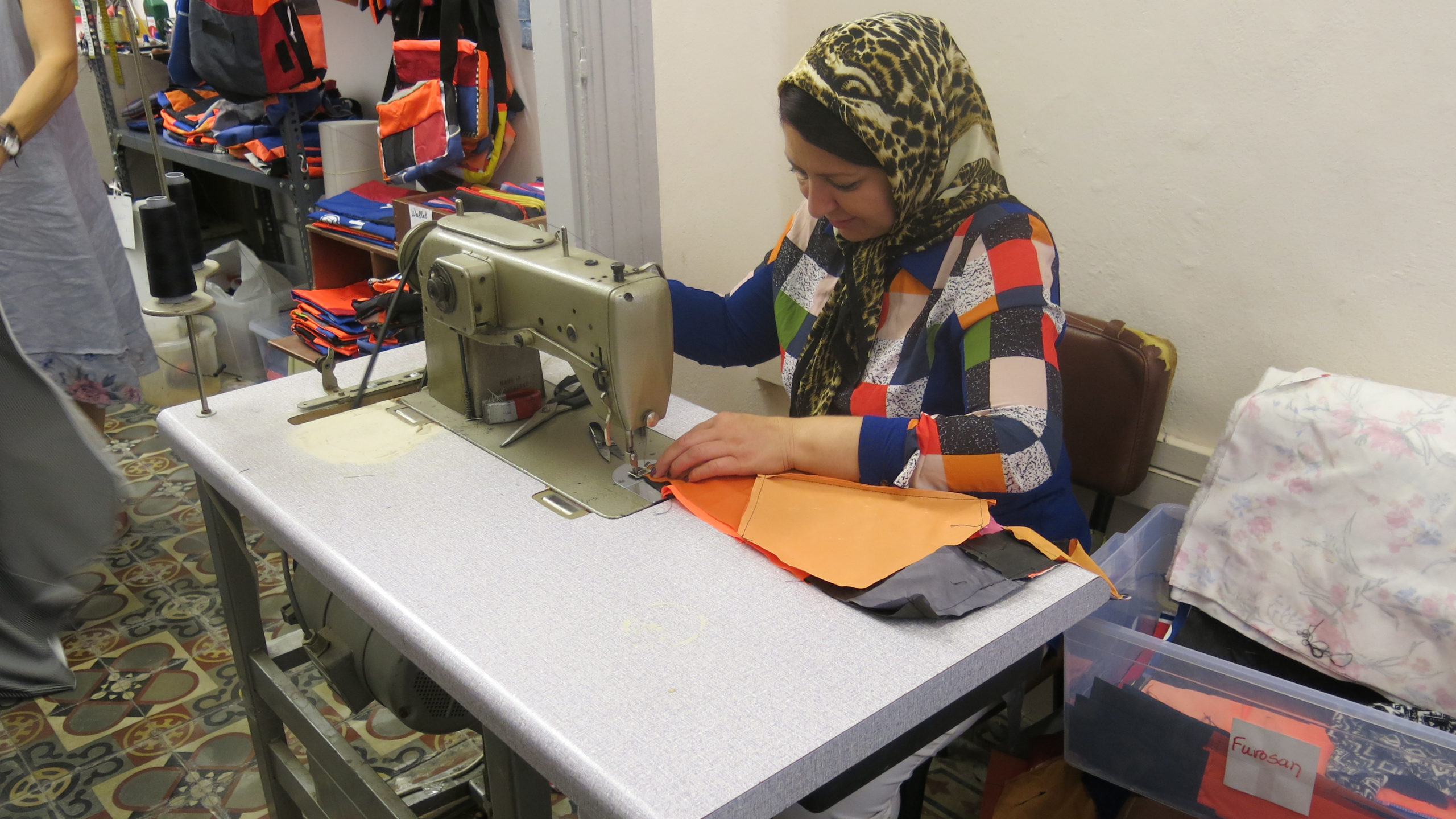 A woman is seated using a sewing machine in a workshop filled with colorful bags.