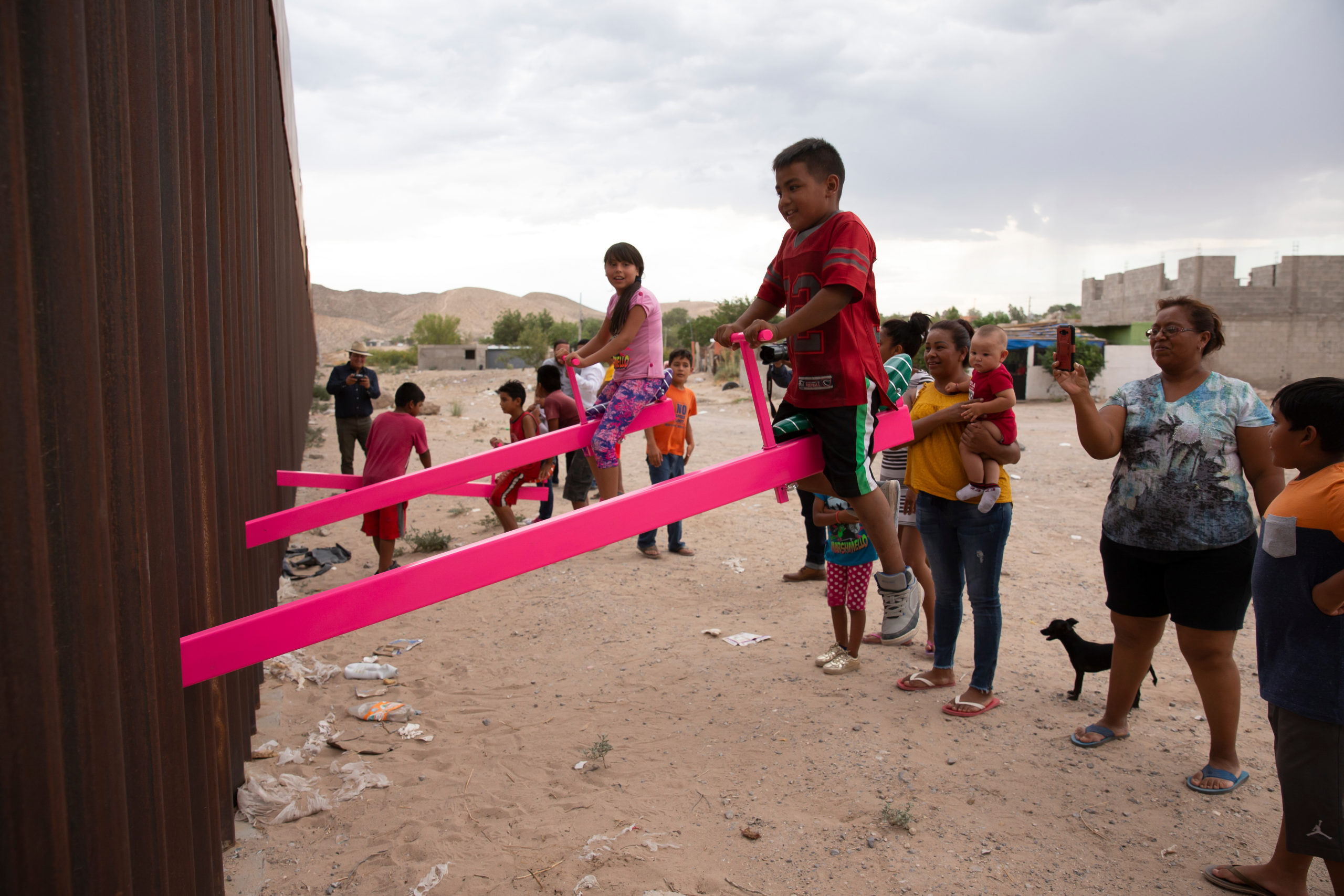 A few brown-skinned children sit and smile on hot pink teeter-totters, the other end of the teeter-totters not visible beyond the tall metal fence that they run through.