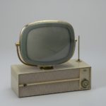 A round-cornered gray television screen suspended on a rounded brass bracket. The bracket is centered on a beige, rectangular platform.