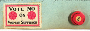 Close up of a section of a blue page on which is affixed a stamp that is outlined in a pink rectangle containing the words "Vote No on Woman Suffrage" beset with two pink flowers. Next to the stamp is a pink circular button that reads "Vote No on Woman Suffrage."