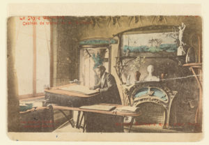 Sepia toned postcard with man sketching at drafting table with ornate fireplace in back. Top of postcard reads "Le Style Guimard"