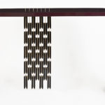 A sleek table consisting of a slender arc joining two legs and a table top. The table is deep red in color and from the table top descends a column of 32 stylized black feathers.