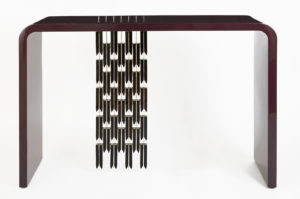 A sleek table consisting of a slender arc joining two legs and a table top. The table is deep red in color and from the table top descends a column of 32 stylized black feathers.