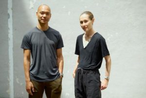 A portrait of a man with medium skin tone and a woman with light skin tone standing in front of a gray wall. The man is bald and wears a gray T-shirt and army green pants. The woman has her hair pulled back and wears a black T-shirt with black pants.