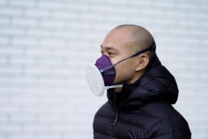 A photograph of a person wearing a black puffer jacket and a purple and white industrial face mask.