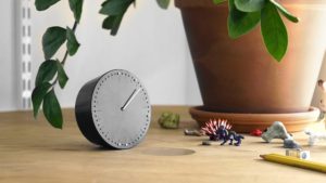 A small, sleek gray clock with tiny holes in place of numbers around the edge of the clock face. It is about a quarter of the size of the flowerpot that sits on the table next to it.