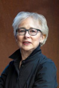 A portrait headshot of a light-skinned woman with short white hair in front of a muted light brown background. She wears black glasses and a black, long sleeve top with the collar up.