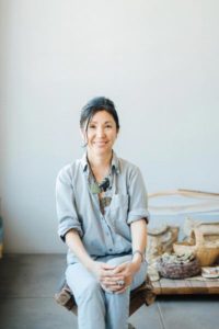 A medium-light skinned woman sits on a stool with her hands placed around her knee. She has black hair pulled up and wears a muted blue jumpsuit and a statement necklace. Behind her are decorative wooden baskets.