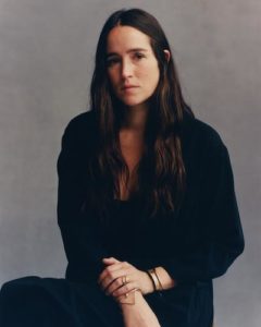 A portrait of a woman with light skin tone sitting with her left hand resting on her right wrist. She has long brown hair and wears a black shirt and black pants with gold bracelets and a gold ring.