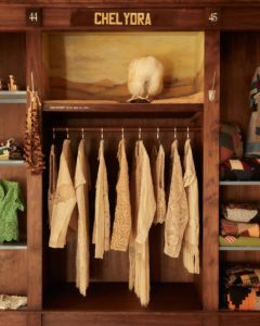 An open wardrobe made of dark wood with clothing in light, neutral tones hanging in the center and thick blankets and other rustic items on the shelves on either side.
