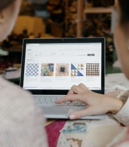 Two people, mostly out of frame, look at several textile patterns on Emily Adams Bode's website on a laptop.