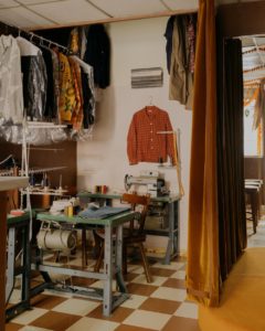 A pair of jeans sits on a table at a small workstation, with clothes hanging on the surrounding walls and a sewing machine behind the table.