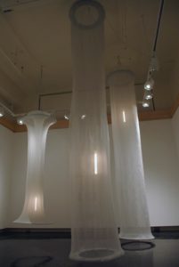 An art installation with three tall cylindrical hanging structures of white mesh-like fabric.