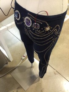 A photograph of the lower half of a mannequin wearing black pants with digital sensors and wires attached.