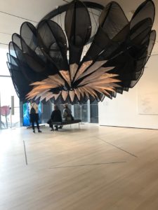 A massive structure made of tubes of black mesh fanned out like a flower hangs from the ceiling in a gallery space.