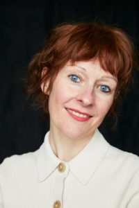 A portrait of a woman with a light skin tone, blue eyes, and red hair in front of a black background. She wears a white button up shirt with tan buttons.
