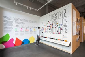 A corner of an exhibition space, with white walls covered in bold black text and colorful geometric shapes. The left wall has a large heading that says [What Shapes Your Choices?] and the right wall has an interactive element titled [Design Your Best Life].