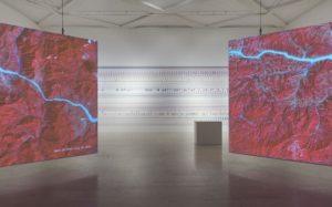 An exhibition space featuring two massive screens showing aerial views of red mountain ranges shot through with the neon blue veins of rivers.