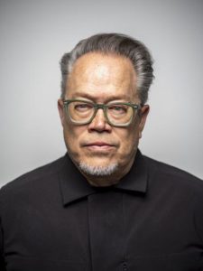 A portrait headshot of a medium-light skinned man looking directly to camera, with short gray hair. He wears a black collared shirt and light green glasses.