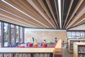 A sun-drenched library filled with low bookshelves and children playing. The ceiling is covered in very thin wooden beams that run parallel along the length of the room.