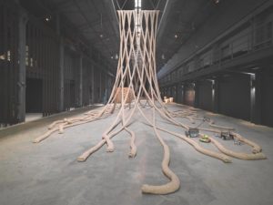 Giant, thick ropes hang down in a crisscross pattern from the ceiling of a large warehouse, their ends curving along the concrete floor like snakes.