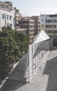 An aerial photograph of a narrow outdoor walkway shaded by a tall, sweeping concrete roof held up by a row of thin pillars.