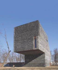 A strangely shaped building made of a textured gray material. The building's top half is a large cube that juts out from the side of the building towards the viewer, with a tall, narrow window in the bottom right corner.