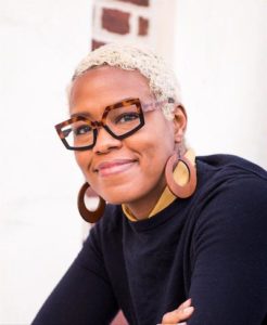 A portrait of a woman with a dark skin tone smiling at the camera. She has short white-blonde hair. She wears a black shirt with a yellow collar, angular brown glasses, and wooden hoop earrings.