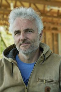 A portrait of a light skin tone man with short white hair and a short gray beard. He wears a light blue T-shirt under a beige jacket.