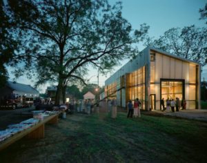 In early evening, people mingle on the grass outside a modern, windowed building lit by a warm, golden glow.