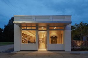 A photograph looking directly into the windowed front entrance of a long, low, rectangular white building labeled [Newbern Library]. The warm yellow glow of the library's well-lit interior contrasts with the cool twilight outside.