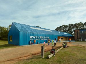 Children gather outside a long, bright blue barn-like building with large white text along the side that reads [Boys and Girls Club. West Alabama Greensboro].