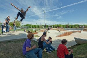 Young people gather at a skate park on a clear, sunny day, with some seated, some standing, and one skater in mid-flight with their arms in the air.