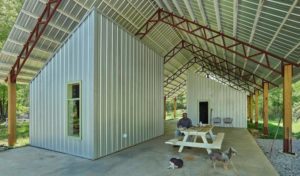 A striking outdoor structure made of corrugated metal, with a high, pointed roof supported by wooden beams covering an open concrete space on which sit two offset enclosed structures.