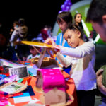 A photograph of a medium-dark skinned child building a structure out of cardboard and paper materials and has a very concentrated look on their face. Craft materials are scattered around the table, and more children are building and creating objects in the background.