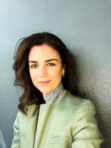 A portrait of a medium-light skinned woman against a textured gray wall. She has medium-long brown hair and wears a muted green jacket with a gray collar.