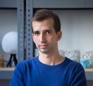 A portrait of a man with a light skin tone looking to camera. He has short brown hair and wears a blue sweater. He sits in front of industrial shelving holding products.