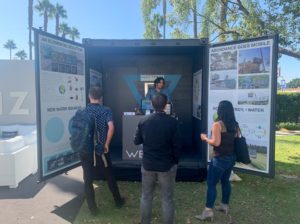 A person stands behind a small kiosk in a shipping container that has been converted to look like a storefront, with the two open doors of the truck covered in informational signs with headings like [An Elemental Solution], [New Water Sources], and [Abundance Goes Mobile].