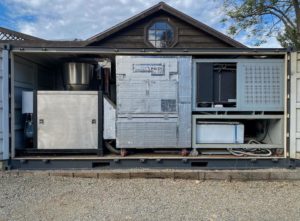 Several pieces of metal machinery inside a portable rectangular structure affixed to the back of a house.
