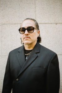 A portrait of a man with medium-light skin tone standing in front of a concrete wall. His hair goes to the top of his shoulders. He wears dark sunglasses, a black blazer that is buttoned up over a black shirt, and a gold necklace.