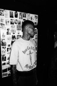 A black and white photograph of a young Black adult with close-cropped hair and a tattooed neck looking to the side and standing in front of a collage of military photographs.