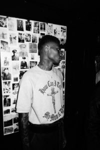 A black and white photograph of a young Black adult with close-cropped hair and a tattooed neck looking to the side and standing in front of a collage of military photographs.