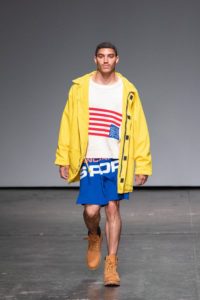 A model with medium brown skin, short, tight, black curls, and a thin mustache walks down a runway in an oversized bright yellow jacket, a white T-shirt printed with an upside down American flag, and baggy blue athletic shorts.
