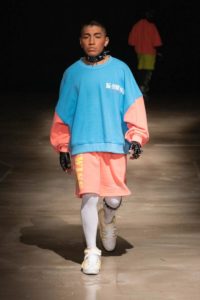 A model with medium brown skin and close-cropped black hair walks down a runway wearing a baggy light blue and salmon pink sweatshirt with matching shorts. They also wear white over-knee socks and spiky black leather fingerless gloves that match their spiky black choker.