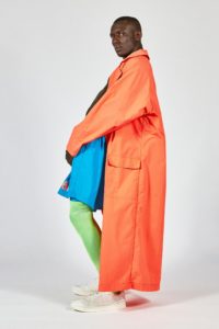 A model with deep brown skin and close-cropped black hair poses in profile, showing off their neon orange floor-length coat, which they wear over a black shirt, bright blue athletic shorts, and neon green above-the-knee socks.