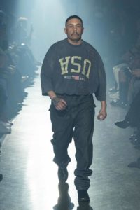 A middle-aged model with tan skin, close-cropped black hair, and a mustache walks down a runway wearing an oversized T-shirt that says USA in upside letters along with baggy jeans.